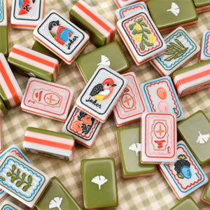 May include: A collection of rectangular mahjong tiles featuring olive green backs and white fronts with colorful engraved illustrations, including joker tiles, floral motifs, and ginkgo leaf designs, scattered across a checkered fabric surface.