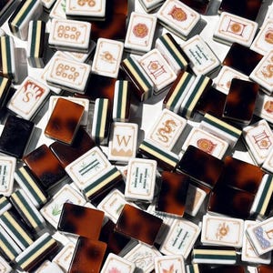 Puede incluir: Una colección de fichas de mahjong rectangulares que presenta una mezcla de fichas blancas con símbolos, caracteres e ilustraciones grabados en rojo, verde y negro, junto con fichas sólidas de color marrón oscuro con patrón de carey.