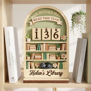 Könnte beinhalten: Holzregal-Display mit dem Text "BOOKS READ THIS YEAR" und der Zahl "136". Das Display enthält Miniaturbücher, Pflanzen und eine schlafende schwarze Katze. Der Name "Helen's Library" ist unten eingraviert. Zwei Bücher befinden sich auf beiden Seiten.