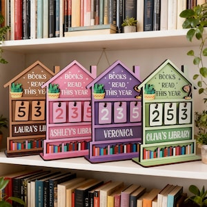 May include: Four wooden book counters in brown, pink, purple, and green, shaped like houses. Each has a cactus, the words "BOOKS READ THIS YEAR", and a name. The counters display numbers, and a shelf of books is visible below.