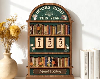 May include: A wooden bookshelf-shaped display with the text "BOOKS READ THIS YEAR." It features shelves filled with miniature books, floral accents, and hanging number tags. The bottom has a plaque that reads "Amanda's Library."