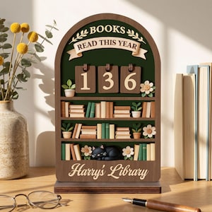 May include: A wooden bookshelf-shaped book counter with the text "BOOKS READ THIS YEAR" and the number "136". The bookshelf has miniature books, plants, and a sleeping black cat. The text "Harry's Library" is at the bottom.