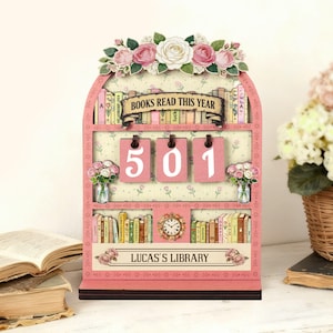May include: A pink wooden bookshelf-shaped desk organiser with the text "BOOKS READ THIS YEAR" and "LUCAS'S LIBRARY". It features a floral design, a clock, and the numbers "501" on hanging tags. Decorative flowers are placed on the top and sides.