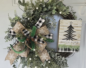 May include: A Christmas wreath with a black and white buffalo check bow, green velvet ribbon, and a wooden sign that says "peace + joy". The wreath is decorated with eucalyptus, white berries, and snowflakes.