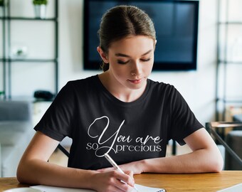 May include: A woman wearing a black t-shirt with the text "You are so precious" in white lettering. She is sitting at a table and writing in a notebook.