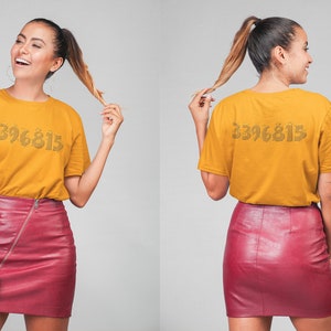 May include: A woman is wearing a mustard yellow t-shirt with the number "3396815" printed on the front. She is also wearing a burgundy leather skirt.