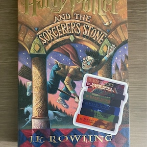 May include: A hardcover copy of "Harry Potter and the Sorcerer's Stone" by J.K. Rowling, featuring a colorful illustration of Harry Potter on a broomstick. A sticker with the titles of the Harry Potter series is on the book.