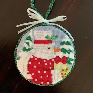 May include: A round, green-trimmed, needlepoint ornament featuring a snowman wearing a red and white hat and a red and white sweater with white polka dots. The snowman is holding a green and white gift box. The ornament is decorated with green and white trees.