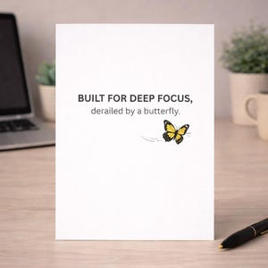 Può includere: Un biglietto bianco con la scritta "BUILT FOR DEEP FOCUS, derailed by a butterfly". Un'illustrazione di farfalla gialla e nera è a destra del testo. Il biglietto è su una superficie di legno chiaro.