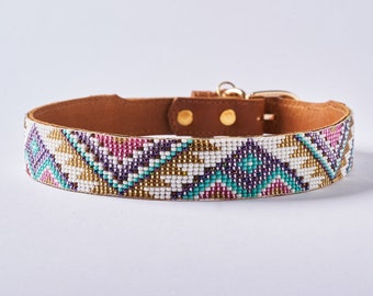May include: A dog collar with a brown leather backing and a beaded front. The beaded section features a geometric pattern in white, teal, gold, purple, and pink. The collar has a gold-colored buckle and rivets.