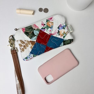May include: A quilted fabric wristlet with a floral patchwork design, a brown wrist strap, and a gold-colored clasp. A pink phone case, lip balm, and coins are also visible. The wristlet has the text "GREEN MANDY".
