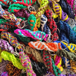 Puede incluir: Una colección de llaveros trenzados de colores con anillos de metal. Los llaveros están hechos de diferentes colores de paracord y están dispuestos en una pila.