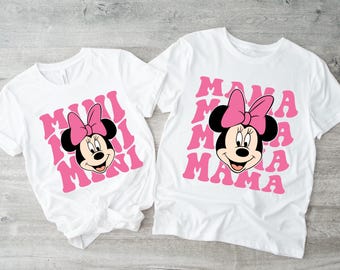 May include: Two white t-shirts with a pink Minnie Mouse graphic. The larger shirt says "MAMA" repeatedly in pink letters. The smaller shirt says "MINI" repeatedly in pink letters.