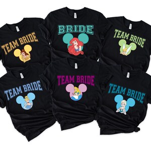May include: Black t-shirts featuring Disney princess designs. Some shirts say "Team Bride" with a princess image inside a Mickey Mouse head. Other shirts say "Bride" with Ariel. The shirts are short-sleeved.