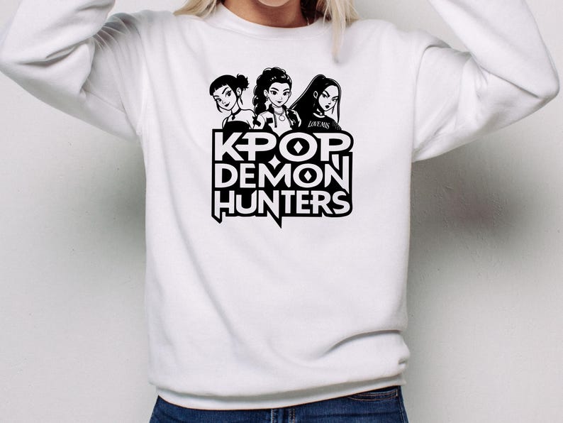 May include: White sweatshirt featuring a black graphic of three stylized female figures above the text "KPOP DEMON HUNTERS." The design is centered on the front of the long-sleeved top. The sweatshirt is made of a soft material.