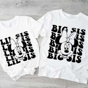 以下が含まれることがあります： 黒いグラフィックの白いTシャツ2枚。1つはミニーマウスのデザインで「Lil Sis」、もう1つはミニーマウスのデザインで「Big Sis」と書かれています。どちらのシャツもレトロなフォントです。