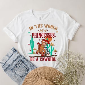 May include: White t-shirt with a graphic print of a cartoon cowboy and horse. The text on the shirt reads "In the world full of princesses be a cowgirl".