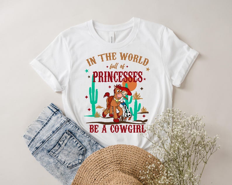 May include: White t-shirt with a graphic print of a cartoon cowboy and horse. The text on the shirt reads "In the world full of princesses be a cowgirl".