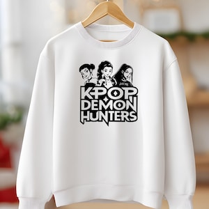 May include: White sweatshirt featuring a black and white graphic of three stylized female figures above the text "KPOP DEMON HUNTERS". The design is centered on the front of the crew neck sweatshirt.
