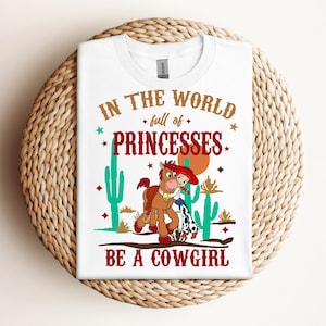 May include: White t-shirt with a brown horse and a girl in a red cowboy hat. The text on the shirt reads "In the world full of princesses be a cowgirl".