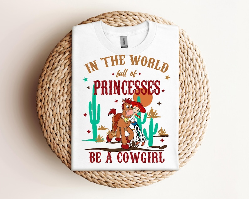 May include: White t-shirt with a brown horse and a girl in a red cowboy hat. The text on the shirt reads "In the world full of princesses be a cowgirl".
