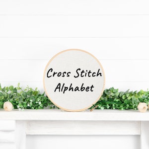 May include: A white embroidery hoop with the words "Cross Stitch Alphabet" written in black on the fabric.