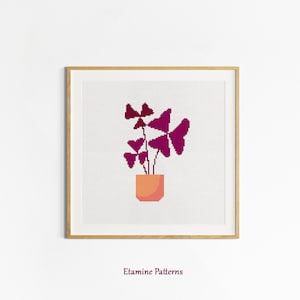 Könnte beinhalten: Kreuzstichmuster einer Topfpflanze mit violetten Blättern. Die Pflanze steht in einem Terrakotta-Topf mit hellorangener Farbe. Das Muster ist in einem hellen Holzrahmen gerahmt.