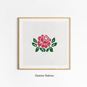May include: A cross-stitch pattern featuring a red rose with green leaves. The pattern is framed in a light wood frame and displayed on a white wall. The text "Etamine Patterns" is printed below the frame.