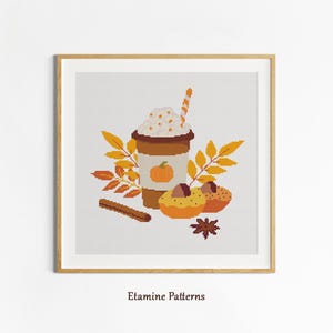 May include: Cross-stitch art of a pumpkin spice latte with whipped cream, a straw, and a pumpkin illustration. The image also includes two muffins, fall leaves, a cinnamon stick, and a star anise. The artwork is framed in a wooden frame.
