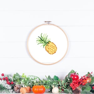 May include: A cross-stitch embroidery hoop with a yellow and green pixelated pineapple design on a white background.