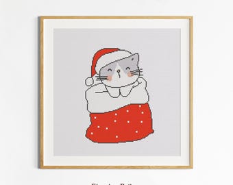 May include: Cross-stitch art print of a cat wearing a Santa hat and sitting in a red and white sack. The cat has a gray face with black whiskers and a pink nose. The print is in a wooden frame, with the text "Etamine Patterns" at the bottom.