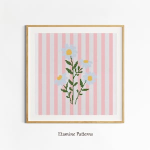 Könnte beinhalten: Ein gerahmtes Kreuzstich-Kunstwerk mit einem floralen Design. Das Kunstwerk zeigt drei weiße Gänseblümchen mit gelben Zentren und grünen Stielen vor einem rosa-weiß gestreiften Hintergrund. Der Rahmen ist hellbraun. Der Text "Etamine Patterns" befindet sich unten.