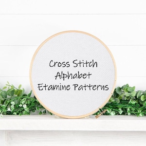 May include: A round embroidery hoop with white cross-stitch fabric. The text "Cross Stitch Alphabet Etamine Patterns" is displayed in black. The hoop is set against a white background with green foliage.