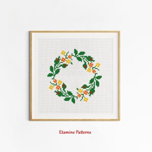 May include: A cross-stitch pattern featuring a wreath of green leaves and yellow and orange flowers. The pattern is framed in a light wood frame. The text "Etamine Patterns" is below the frame.