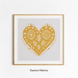 May include: A framed cross-stitch pattern featuring a yellow heart design with sun and leaf motifs. The heart is centered on a white background, and the frame is a light wood color. The text "Etamine Patterns" is at the bottom.