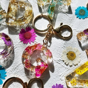 May include: A set of resin letter charms with gold flakes and pressed flowers. The charms are attached to gold key rings with a lobster clasp. The charms are in the shape of the letters D, G, and K. The charms are a variety of colors, including pink, blue, green, and yellow.