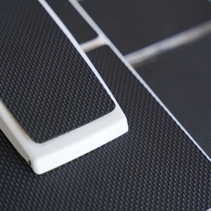 Puede incluir: Primer plano de un pedal de pie texturizado blanco y negro con un borde blanco. El pedal está hecho de un material duradero y tiene una superficie texturizada para un mejor agarre.
