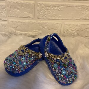Puede incluir: Un par de zuecos azules brillantes adornados con una densa disposición de cuentas multicolores, perlas y pequeños acentos metálicos. El calzado presenta un adorno decorativo de color dorado en forma de lazo sobre cada zapato.
