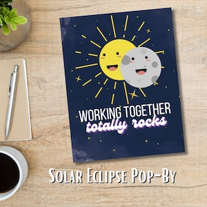 May include: A blue card with a yellow sun and a gray moon with smiling faces. The sun and moon are partially overlapping, creating a solar eclipse. The text "WORKING TOGETHER totally rocks" is written in purple on the card. The card is labeled "SOLAR ECLIPSE POP-BY" at the bottom.