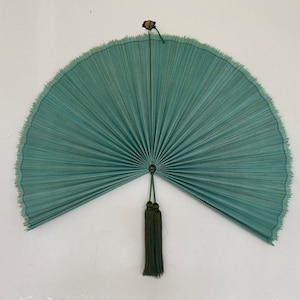 May include: A teal fan with a semi-circular shape, featuring a central circular design and a dark green tassel. The fan's edges have a fringed texture, and it is mounted on a white wall.