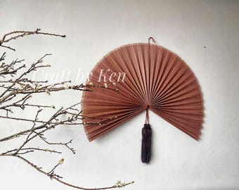 May include: A large, brown, hand-woven fan with a tassel hanging from the bottom. The fan is made of natural materials and has a rustic, bohemian style.