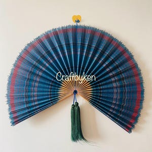 Può includere: Un grande ventaglio aperto con un motivo a quadri blu, rossi e grigi. Il ventaglio è tenuto insieme da uno spago blu e ha un nappa verde. Il testo "craftbyken" è visibile sul ventaglio.