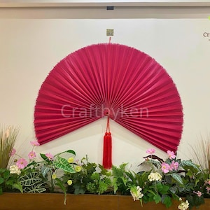 Può includere: Un grande ventaglio da parete a forma di semicerchio, color fucsia, con un nappa, fissato contro un muro neutro. Il testo "CraftbyKen" è visibile. Decorazione sopra piante e fiori.