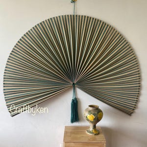 Può includere: Un grande ventaglio da parete a forma di semicerchio con strisce di bambù verde acqua e naturali alternate. Il ventaglio è adornato da un nappa verde acqua ed è montato su una parete. Un vaso decorativo con disegni floreali gialli è posto su una superficie di legno.