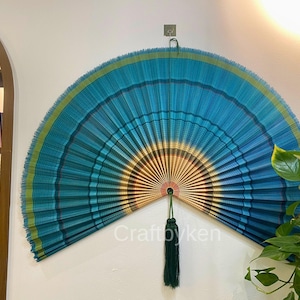 Può includere: Grande ventaglio decorativo nei toni del blu, verde acqua e oro, montato su una parete bianca. Il ventaglio ha un design a pieghe e un nappa verde scuro. Il testo "Craftbyken" è visibile sul ventaglio.