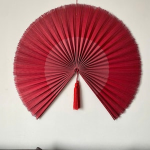 May include: A large red and pink woven fan with a tassel hanging from the bottom. The fan is hanging on a white wall.