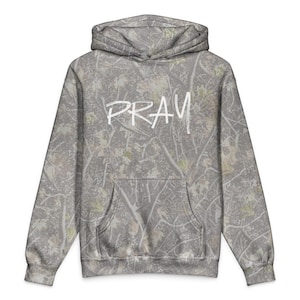 Peut inclure: Un sweat à capuche gris camouflage avec le mot "PRAY" en écriture blanche sur la poitrine. Le sweat à capuche a une poche avant et une capuche. Le tissu présente un motif de branches et de feuilles.