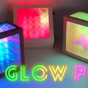 Könnte beinhalten: Drei beleuchtete, würfelförmige Lampen mit farbenfrohen Lichtanzeigen. Eine leuchtet blau, eine rot und die dritte grün. Der Text "GLOW POP" wird in Neonfarben am unteren Bildrand angezeigt.