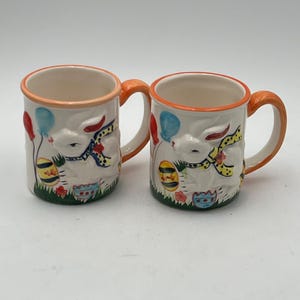 May include: Two white ceramic mugs with orange rims. Each mug features a colorful illustration of a bunny holding an Easter egg with balloons in the background.