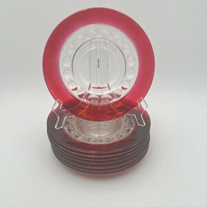 Puede incluir: Una pila de ocho platos de postre de vidrio transparente con un borde rojo. Los platos tienen un patrón elevado de pequeños círculos alrededor del borde.